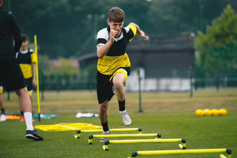 Young man ina yellow and balck top at a sports event running over spaced poles, short hurdles on a field.